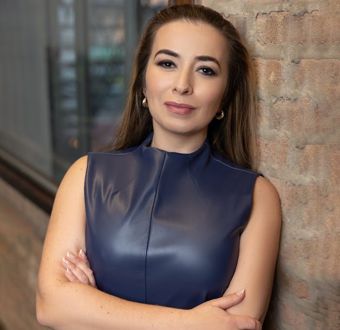 A woman with long brown hair in a sleeveless navy blue top stands with arms crossed, leaning against a brick wall, looking at the camera with a neutral expression—much like the resolve of Chicago immigration attorneys.