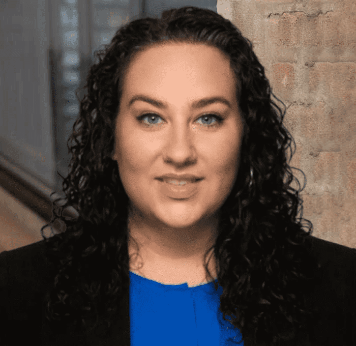 A person with long curly dark hair and blue eyes, wearing a blue top and black blazer, stands indoors next to a brick wall.