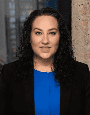 A woman with long curly dark hair, wearing a blue top and black blazer, poses in front of a brick wall and blurred indoor background.