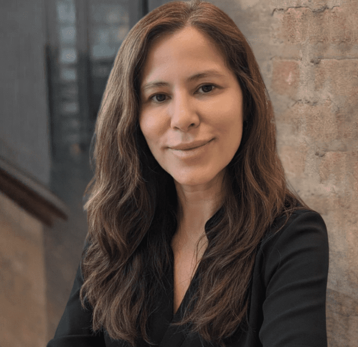 A woman with long brown hair and a black top sits in front of a brick wall, smiling slightly at the camera.